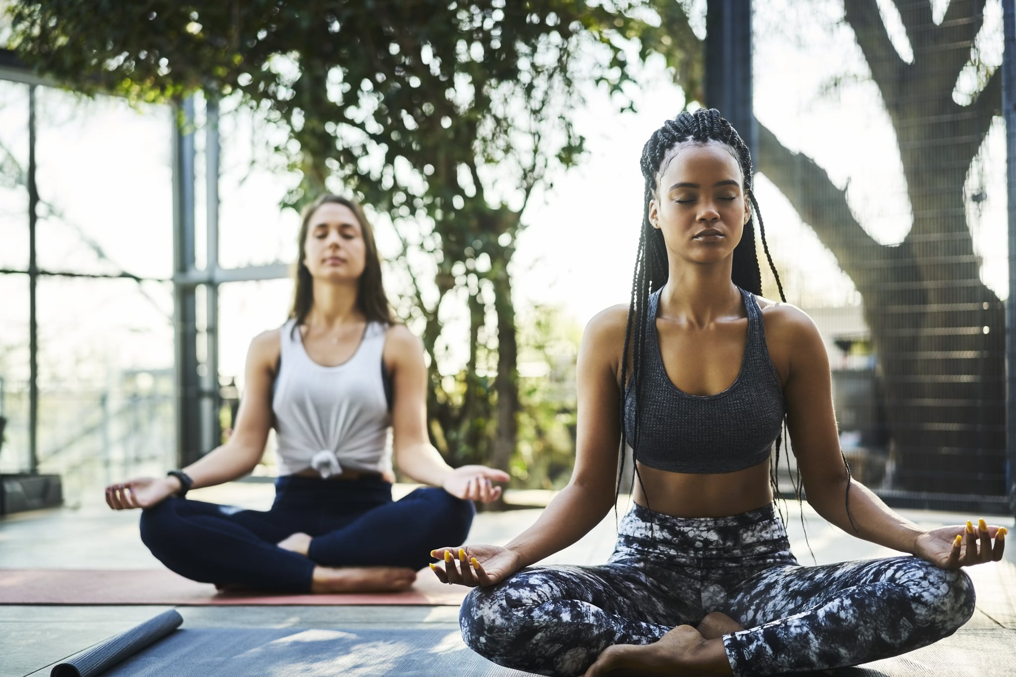 Young woman practicing lotus position with friend on porch. Multi-ethnic females are meditating in yoga class. They are in sports clothing.