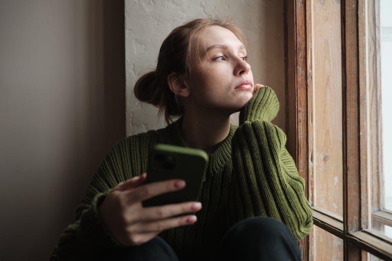 Woman holding phone sitting on windowsill, thinking about trying a sobriety app.