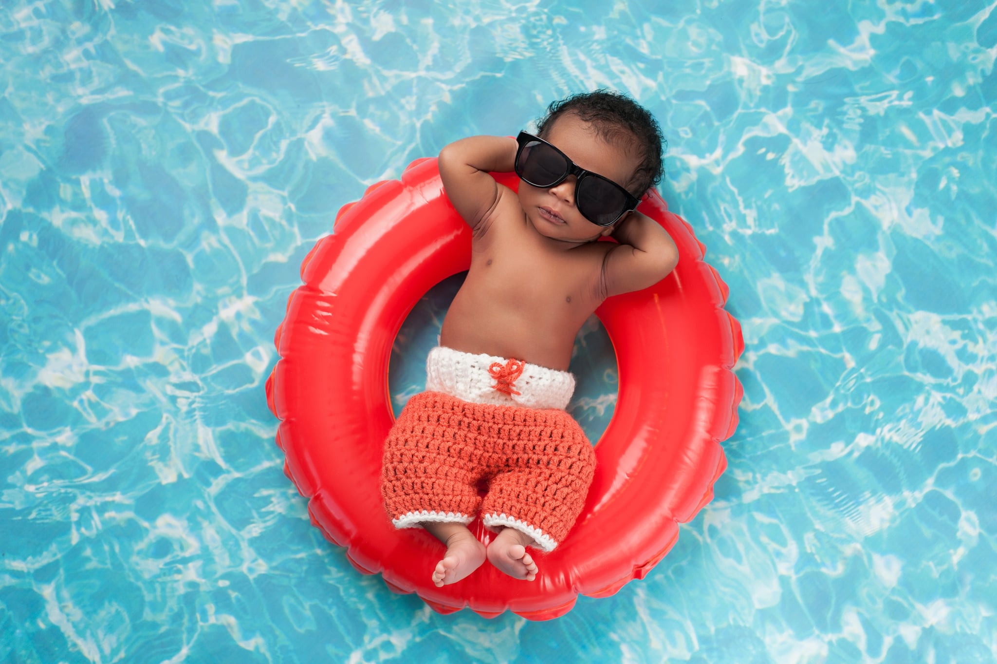 Two week old newborn baby boy sleeping on a tiny inflatable swim ring. He is wearing crocheted board shorts and black sunglasses.