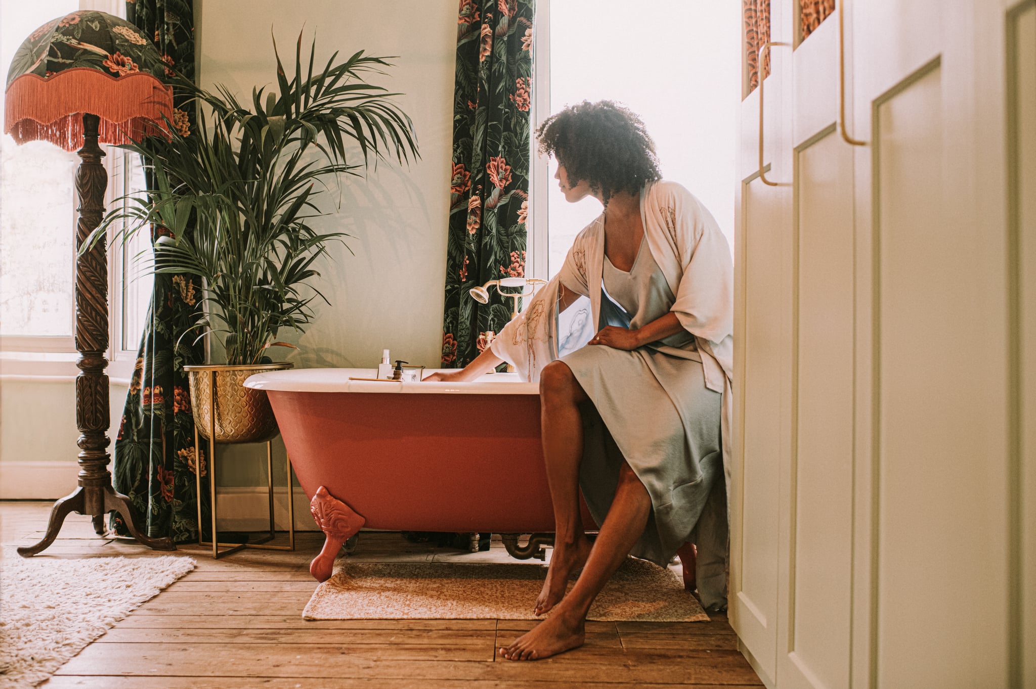 A beautiful woman perches on the side of an elegant red roll top bathtub, and waits for it to fill up. She wears a silky robe. Light floods through the window, backlighting the relaxing scene and giving it a dreamy vibe.