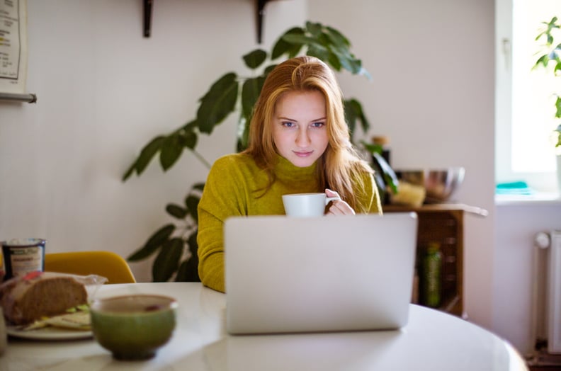 Young woman sitting at table looking at her laptop and drinking coffee. Female using laptop at home.