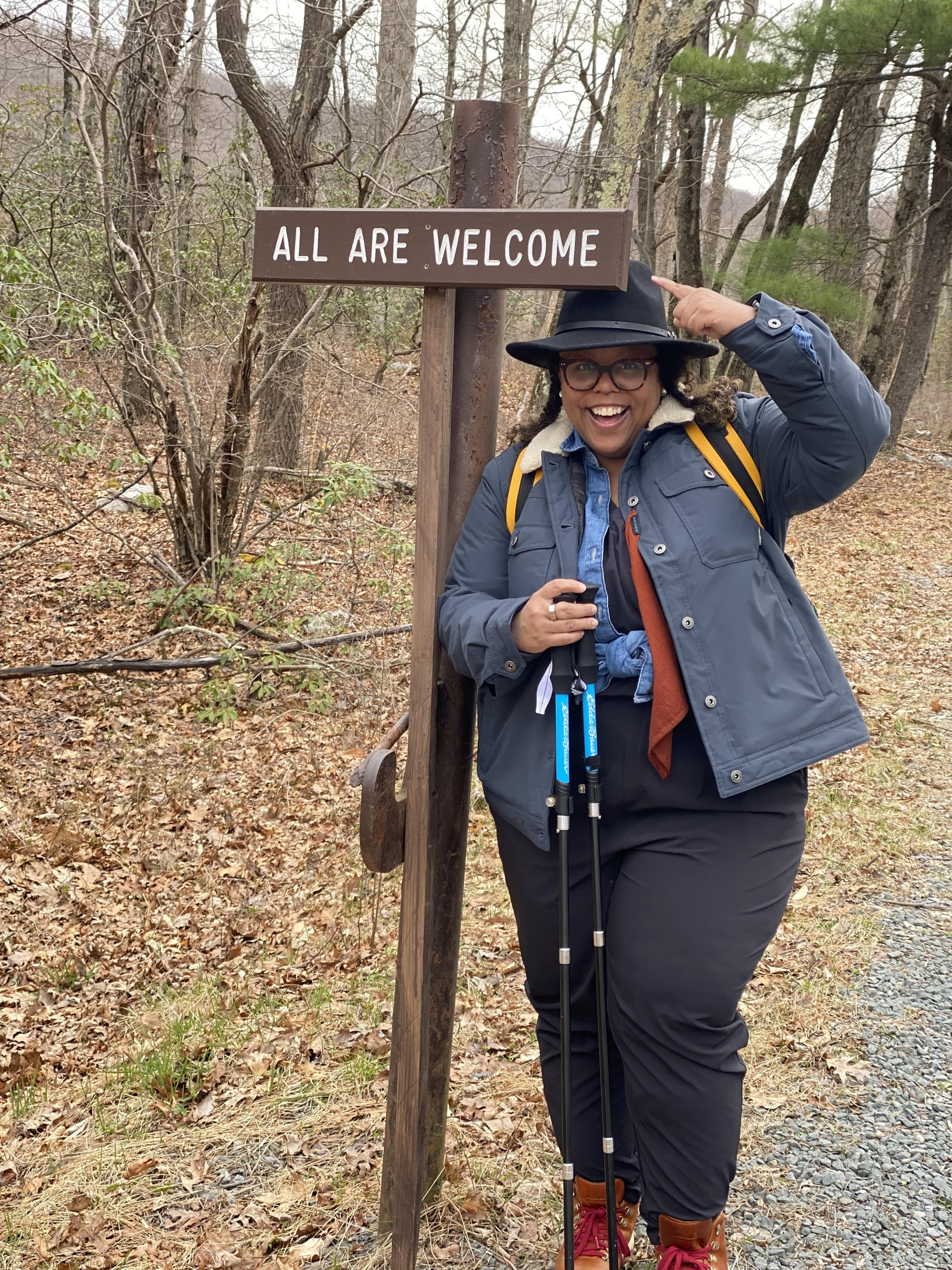 Kisha Jarrett, a Black woman, posing by an "All Are Welcome" sign while hiking the Pacific Northwest trail. Jarrett is wearing Black pants, tan hiking boots, a grey-blue jacket, and black hat.