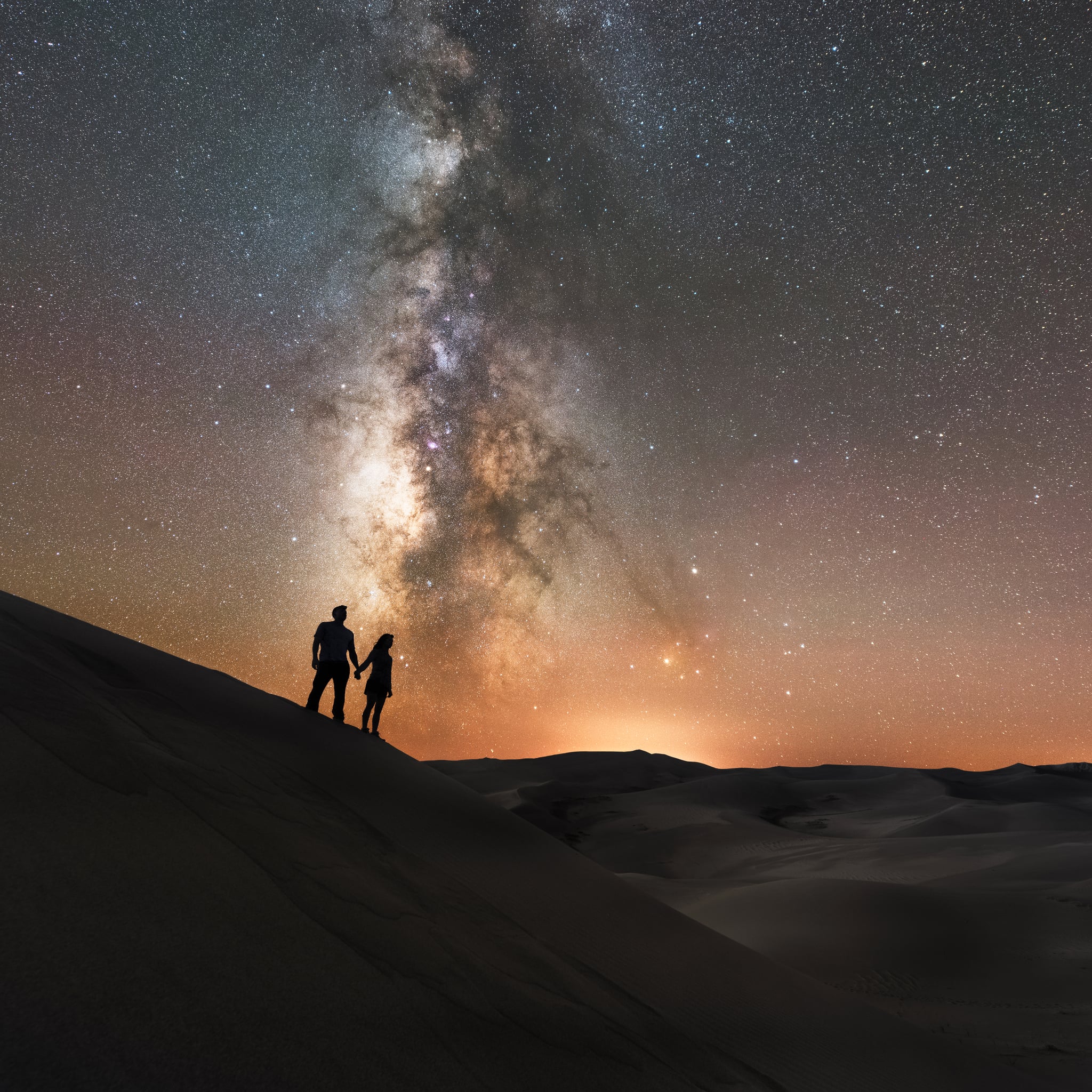 Hikers reach the peak of a sand dune and hold hands under the night sky.
