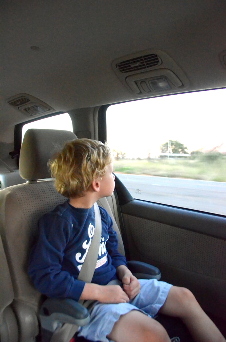 A young boy sits in a booster seat looking out the window of a moving vehicle.