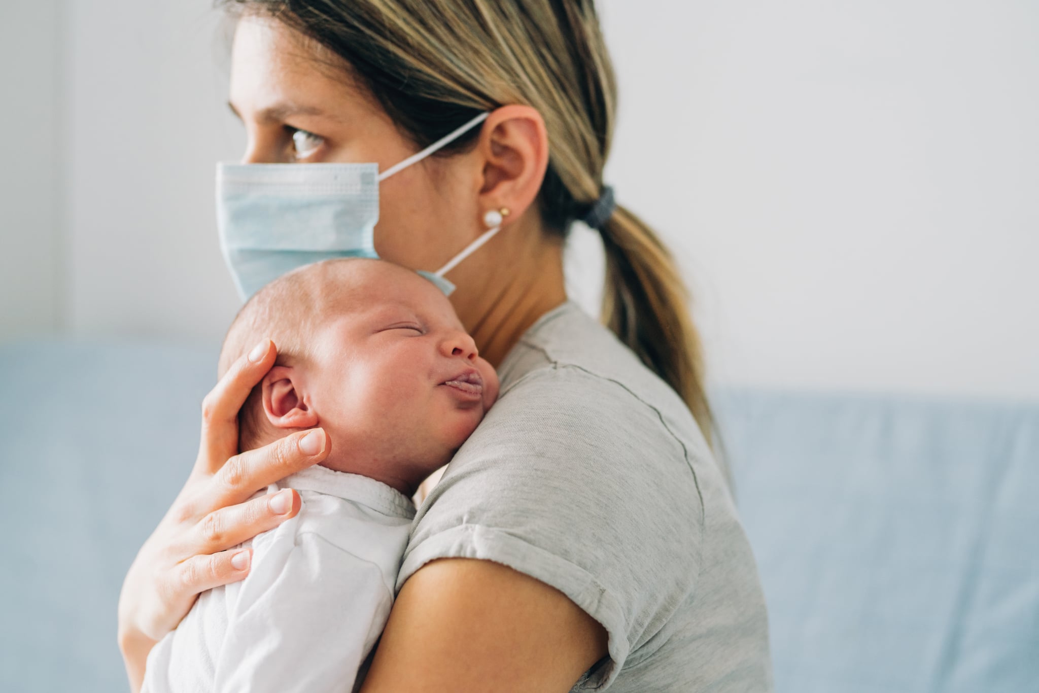 Young mother with protective mask embracing  her newborn baby girl while they are in home isolation during coronavirus/COVID-19 quarantine.