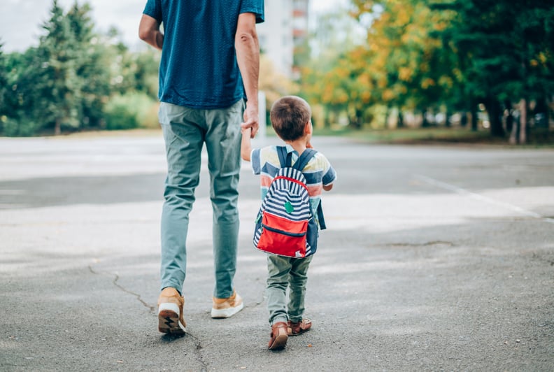 Rear view of father who leads a little boy hand in hand to kindergarten. Father and son with backpack walking in schoolyard.