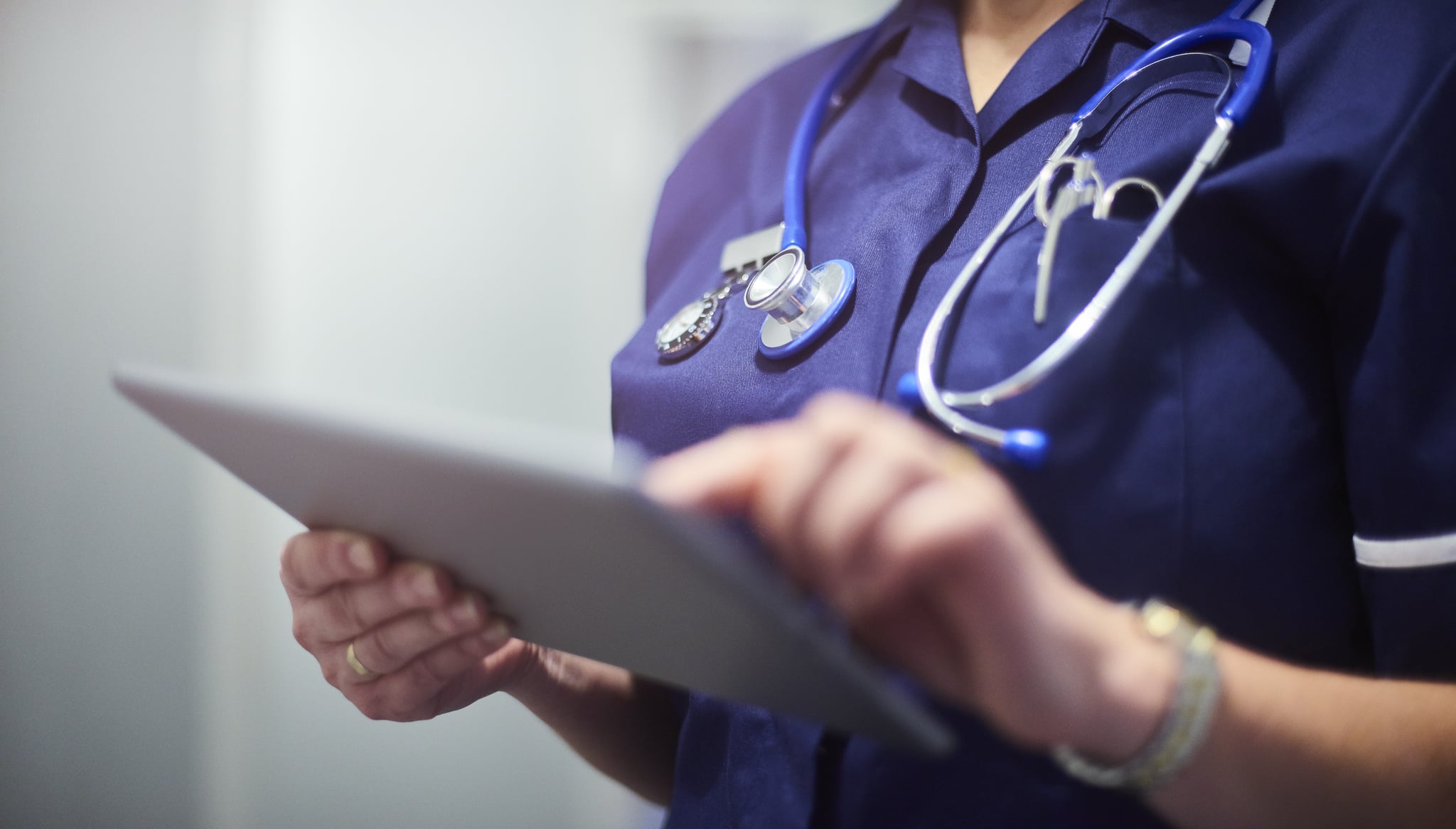 Female surgeon typing on digital tablet in hospital or surgery. She is wearing a dark blue nurse's top and has her stethoscope around her neck. She is looking at her patients records on her digital tablet and sending emails.