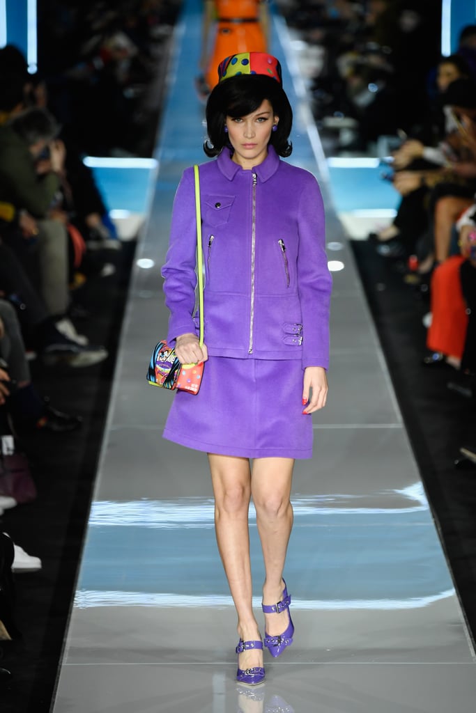 Walking in Moschino wearing a purple outfit and colorful pillbox hat.