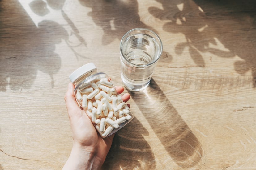 Woman holding clear bottle with white capsule pills beside a glass of water on a wooden counter. 