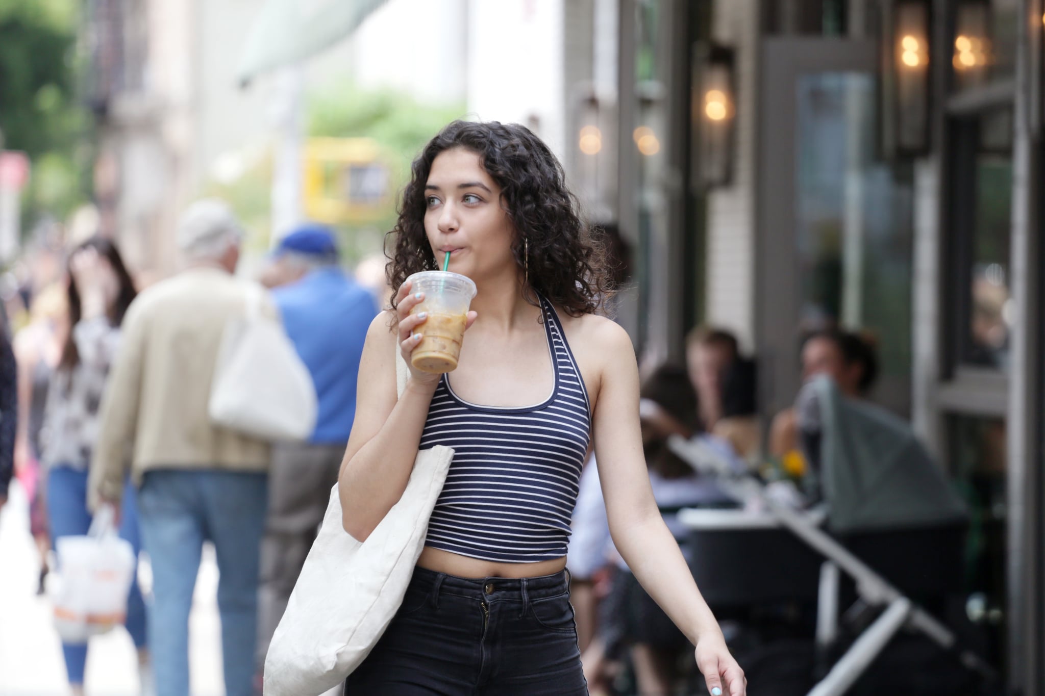 A young LatinX woman wearing a blue and white striped top, with curly hair, happily walks down a New York City street holding an iced coffee.