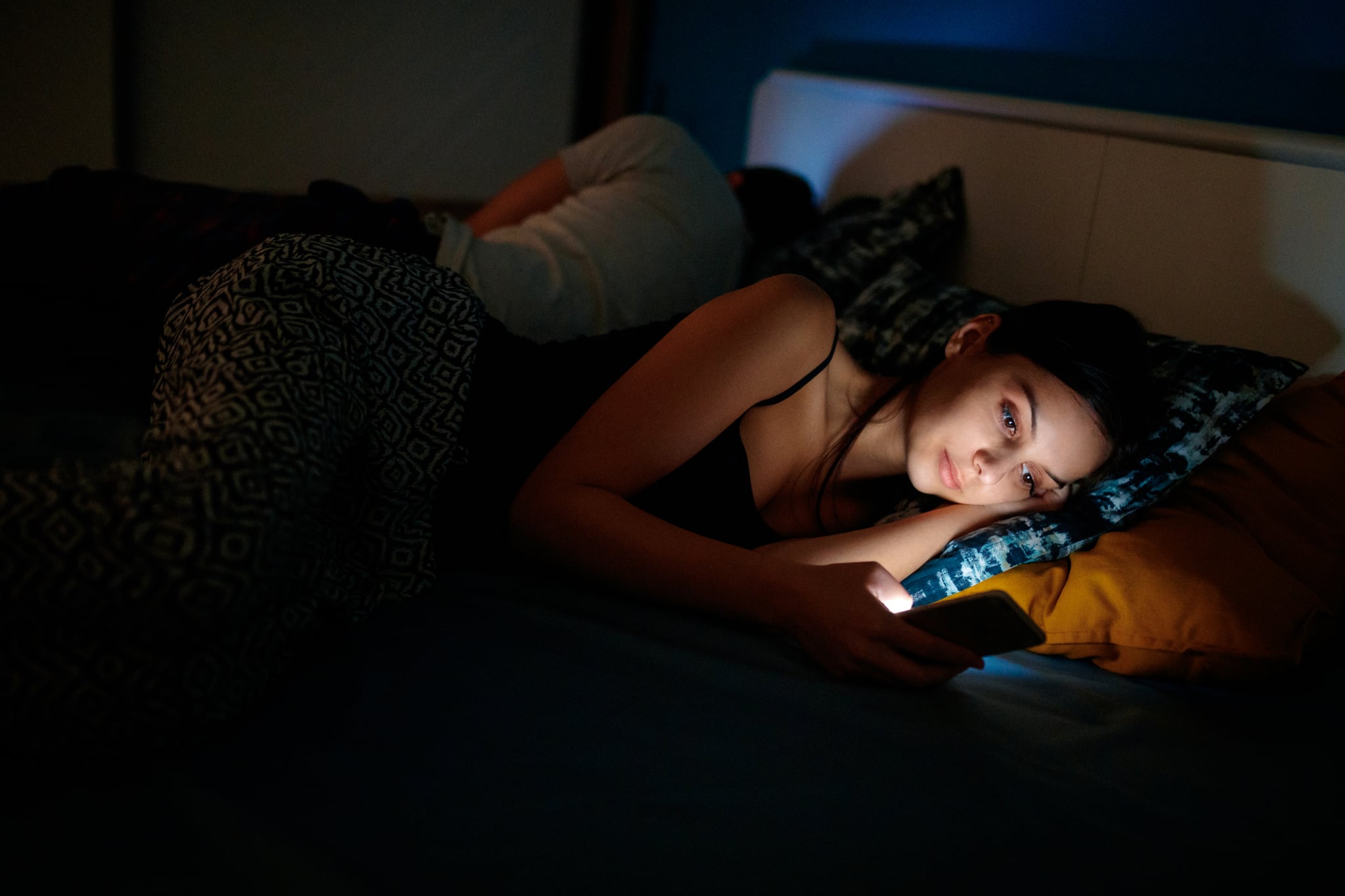 Young woman sending text message as her boyfriend sleeps in the bed, dark, copy space