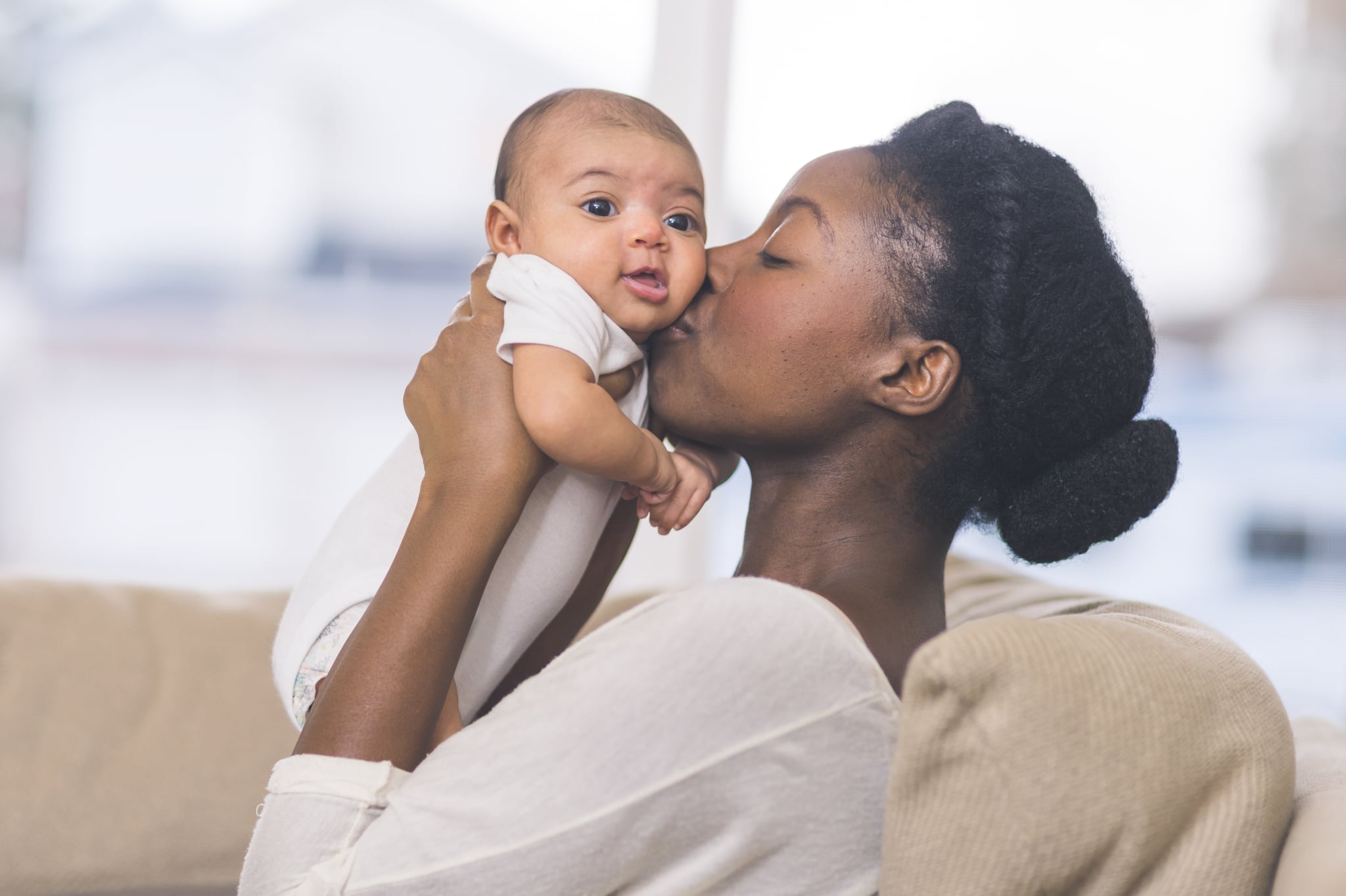 一个美丽的年轻的非洲裔美国母亲温柔地抱着她婴儿的女儿双手在空中和亲吻她的脸颊。婴儿的眼睛睁大,她看上去很幸福。他们坐在客厅的沙发上。