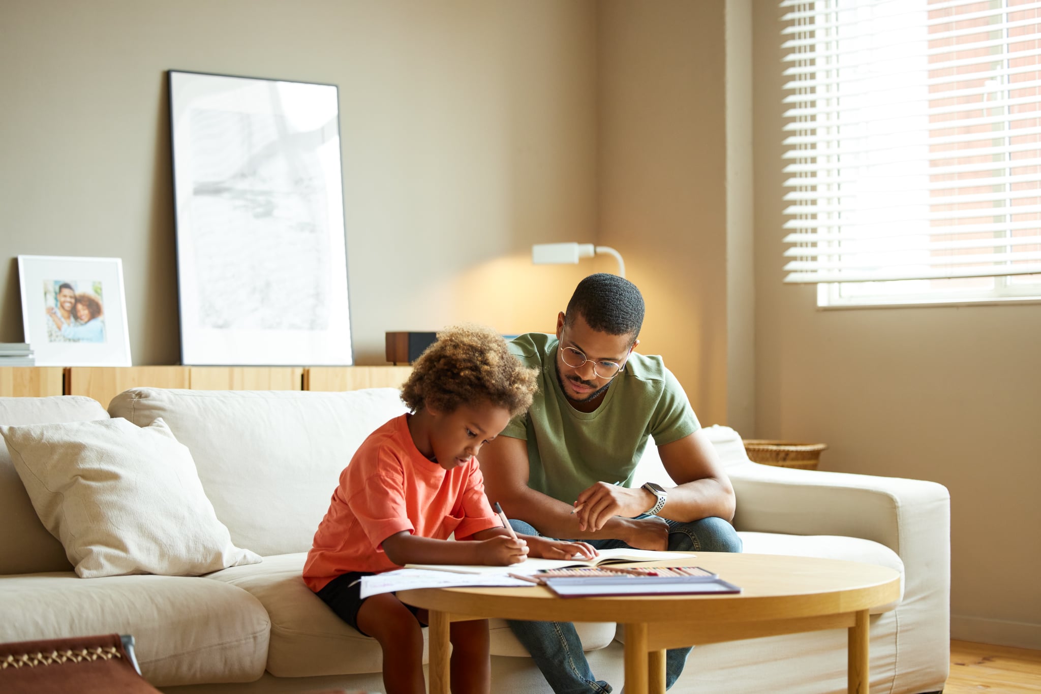 Man wearing eyeglasses sitting by son doing homework at coffee table. Afro boy is writing in book by father. They are in living room at home.