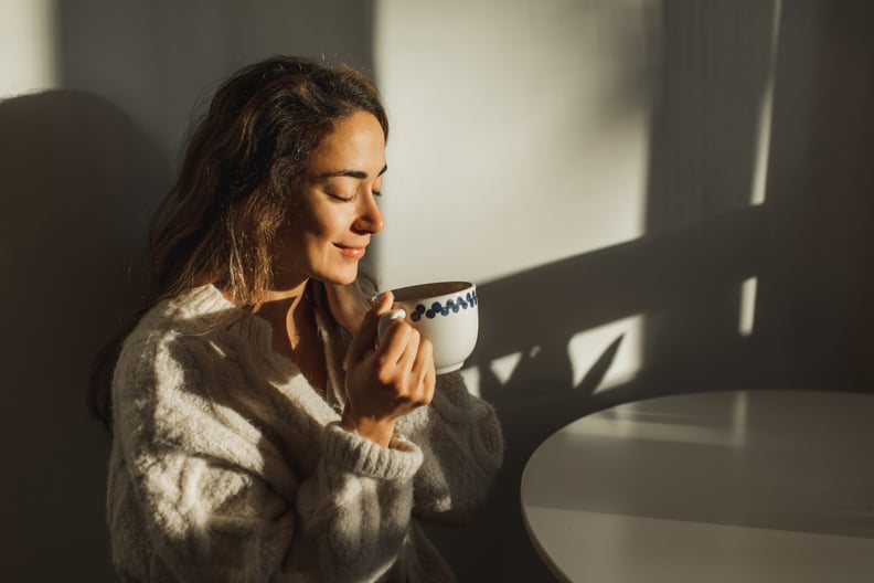 A woman in a big cozy sweater drinking her morning coffee from a mug at the kitchen table.