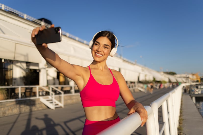Happy young adult sportswoman taking a break from her outdoor training session, having her wireless headphones on and taking a selfie using a smart phone