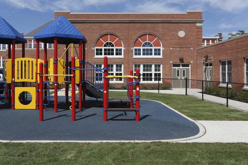 gated elementary school playground with colorful jungle gym equipment, slides, monkey bars, mazes, on padded play surface, with large brick school building complex