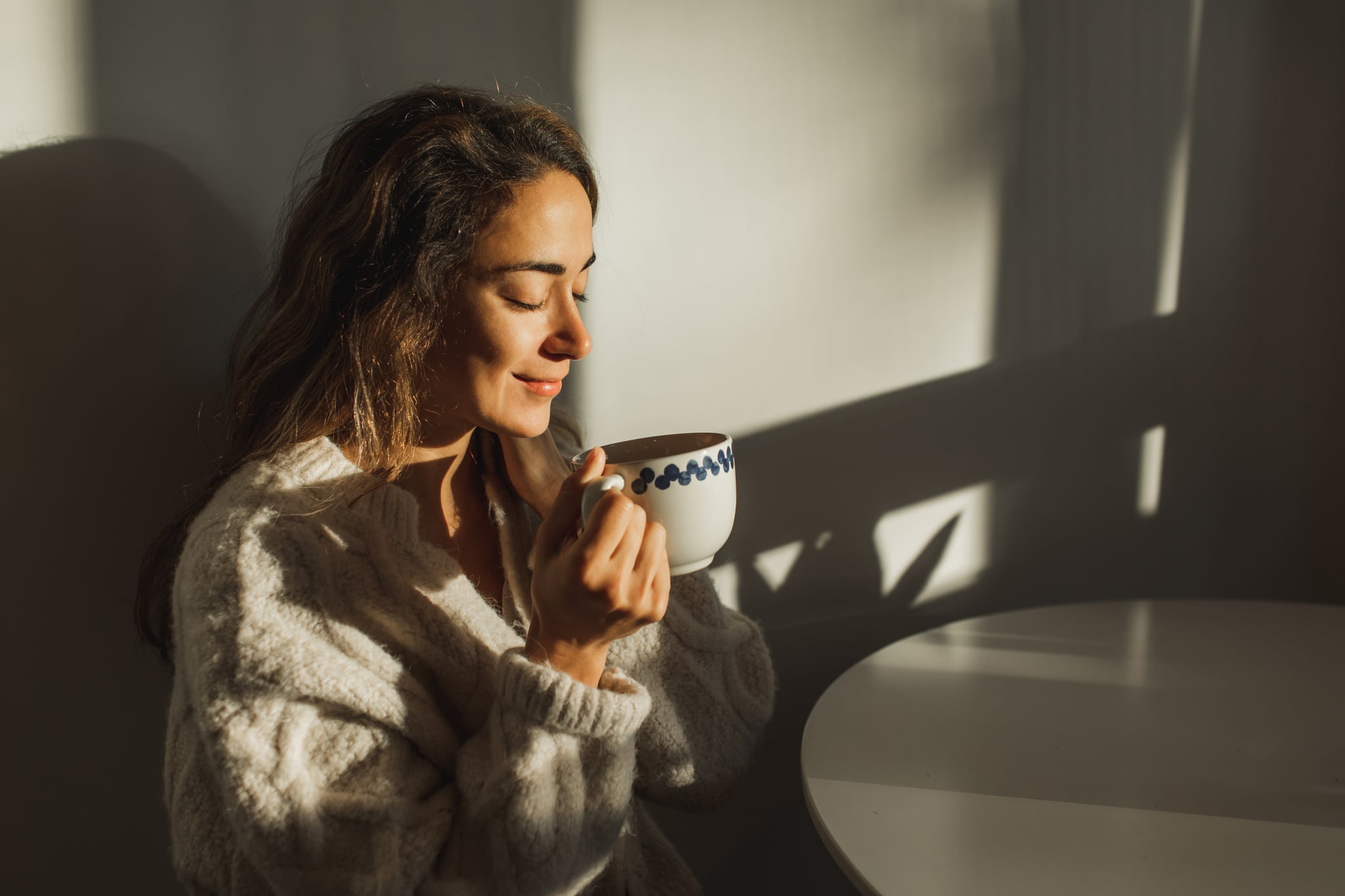 A woman in a big cozy sweater drinking her morning coffee from a mug at the kitchen table.