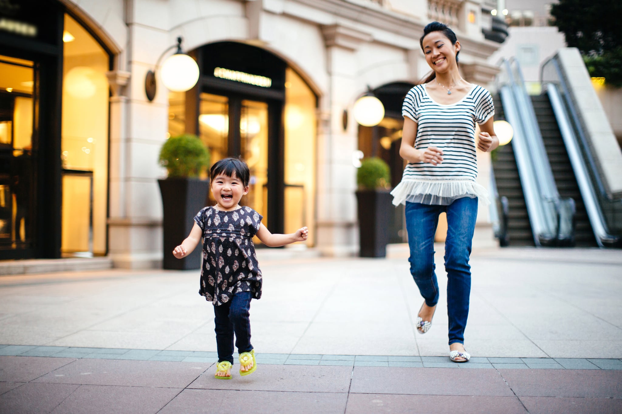 Pretty young mom playing with and chasing after running toddler girl in the square, both smiling joyfully.