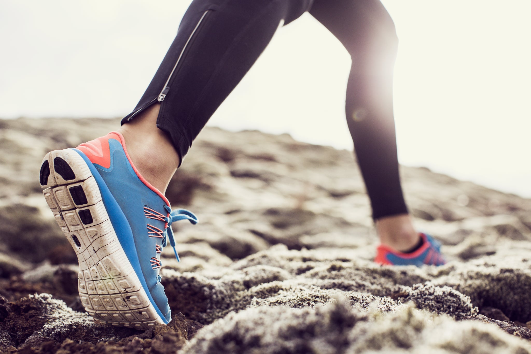 Close up, low angle shot of a female runners trainers, running through mossy terrain in Iceland. Blue trainers, black running leggings with backlit sunshine.