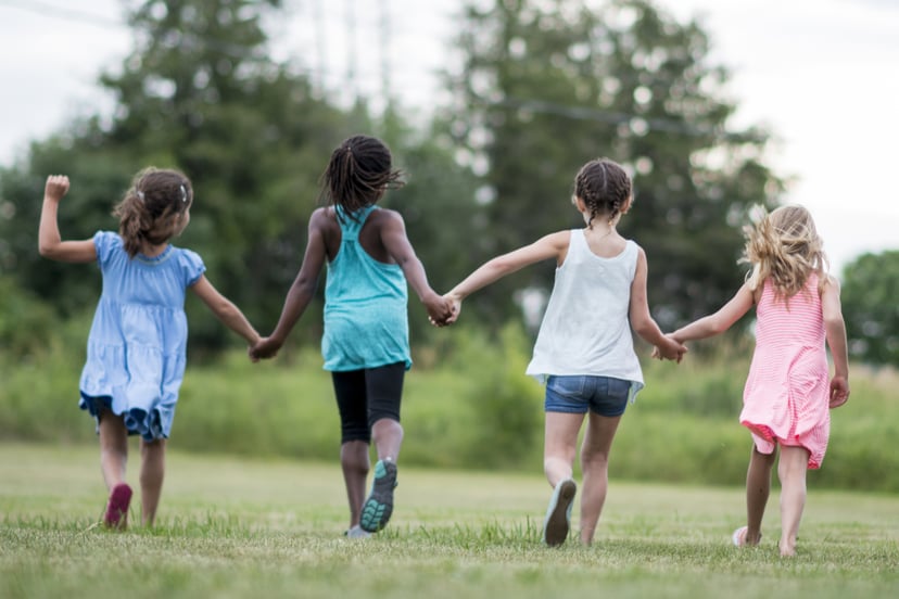 A multi-ethnic group of elementary age children are playing together outside at recess. A group of girls are holding hands and running together in a row.