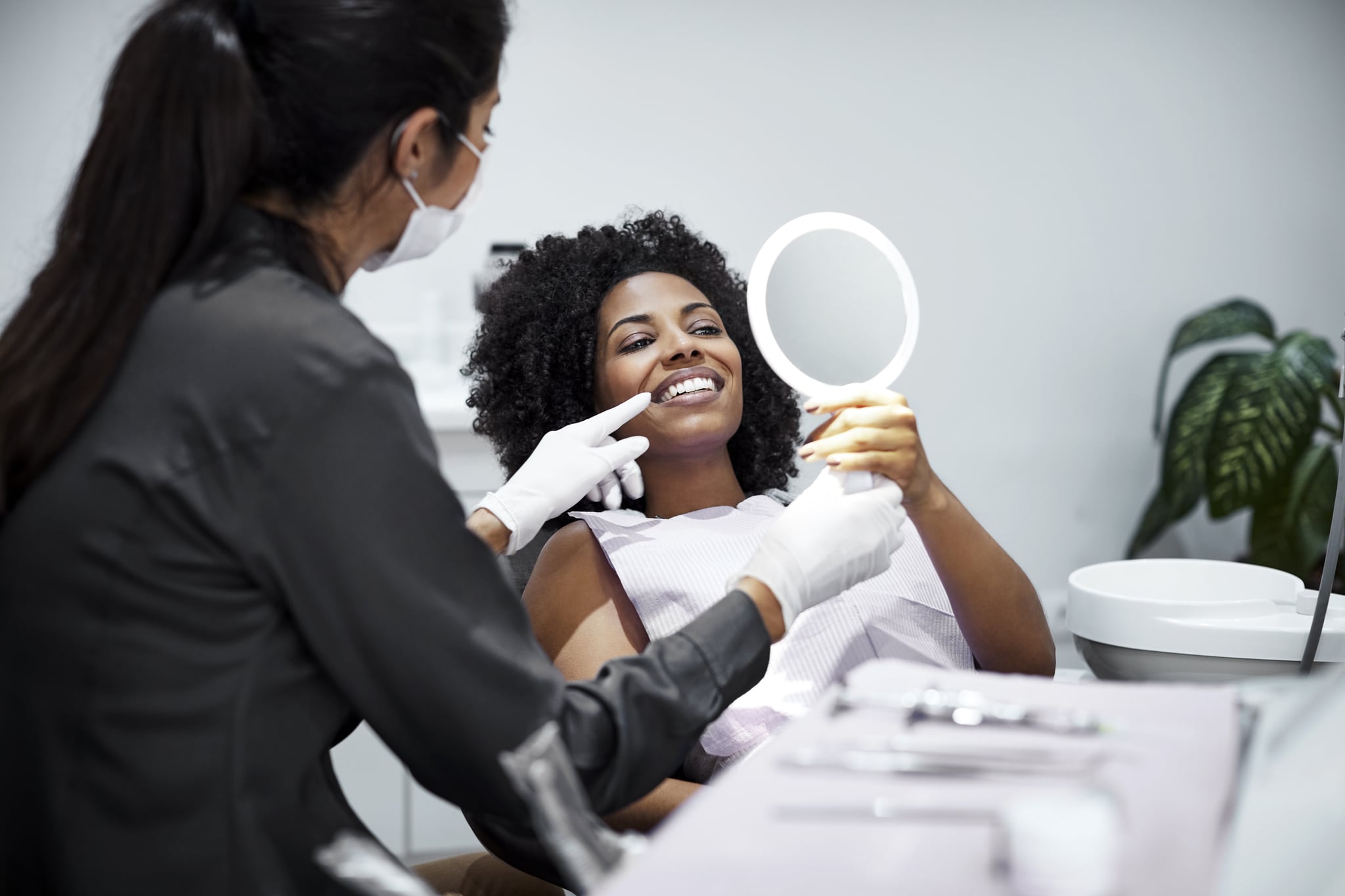 Smiling woman looking teeth in hand mirror. Female dentist is explaining dental health to patient. They are in medical examination room.