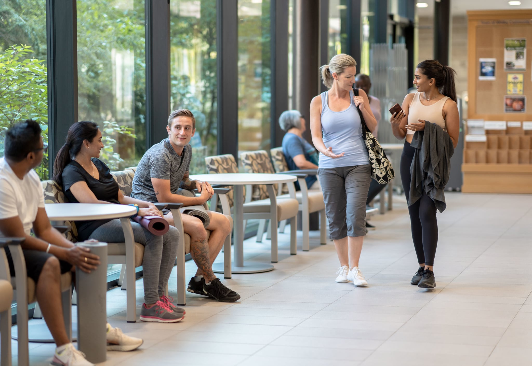 Two women walk through the annex of a gym and joyfully greet the people they see.