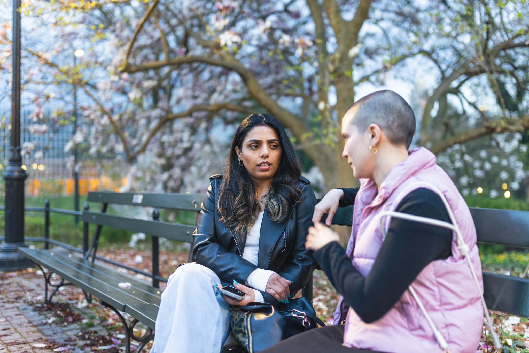 Two women friends having a deep conversation about spring cleaning their friendships on a park bench surrounded by blossoming trees during the evening