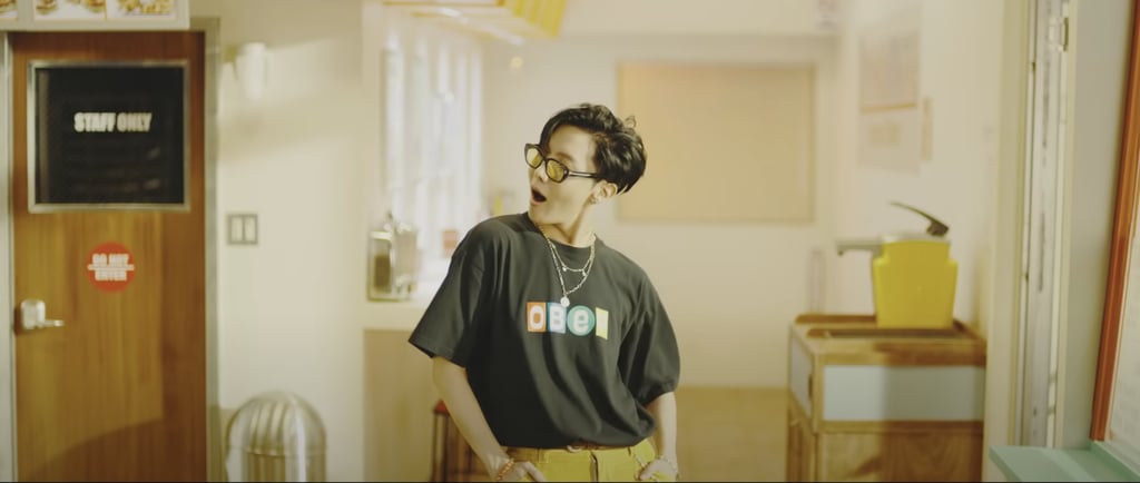 J-Hope in a graphic Obey T-shirt, yellow pants, layered necklaces, and yellow sunglasses is a look we are very much here for.