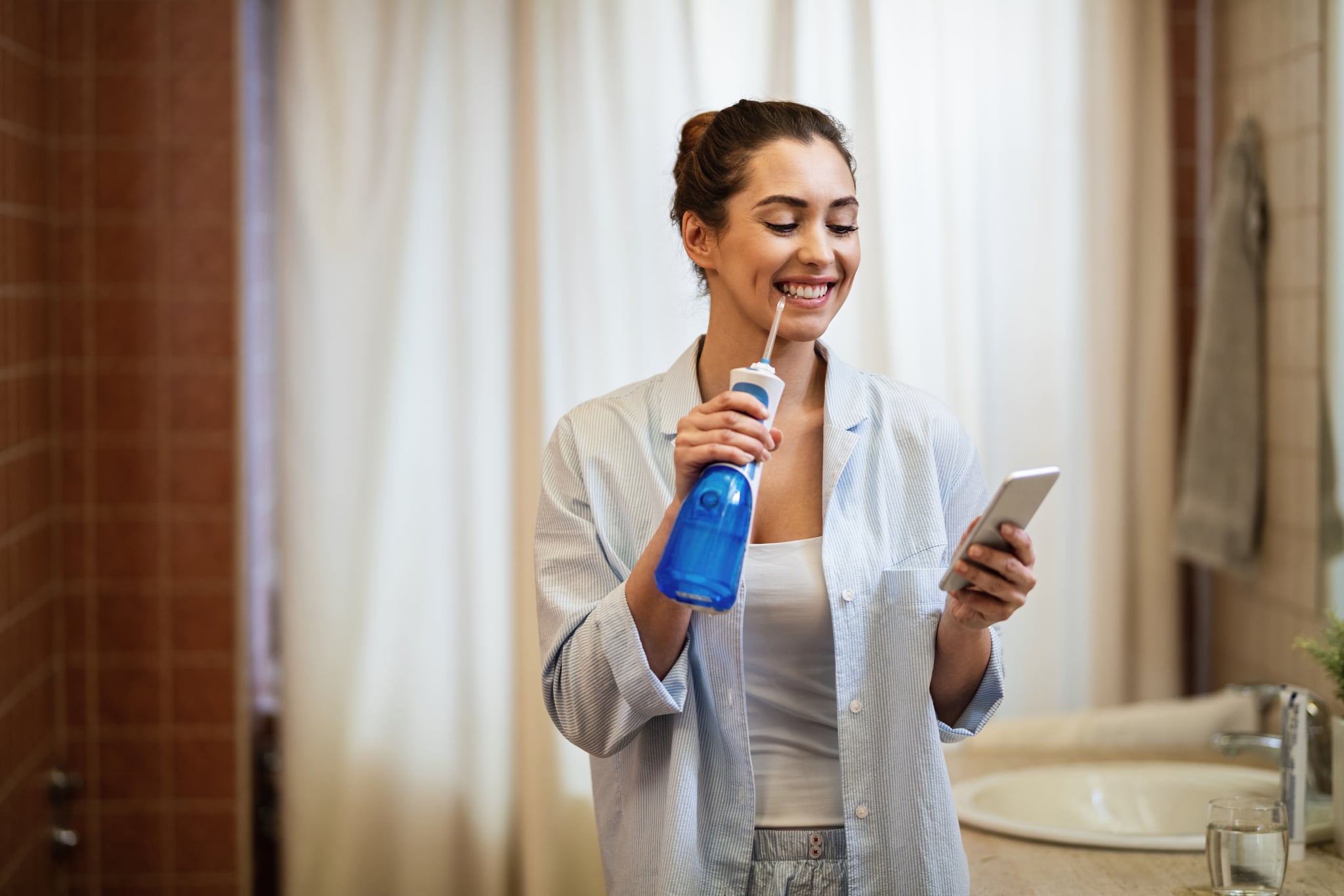 Young happy woman using dental water flosser and cleaning her teeth while texting on mobile phone in the bathroom.