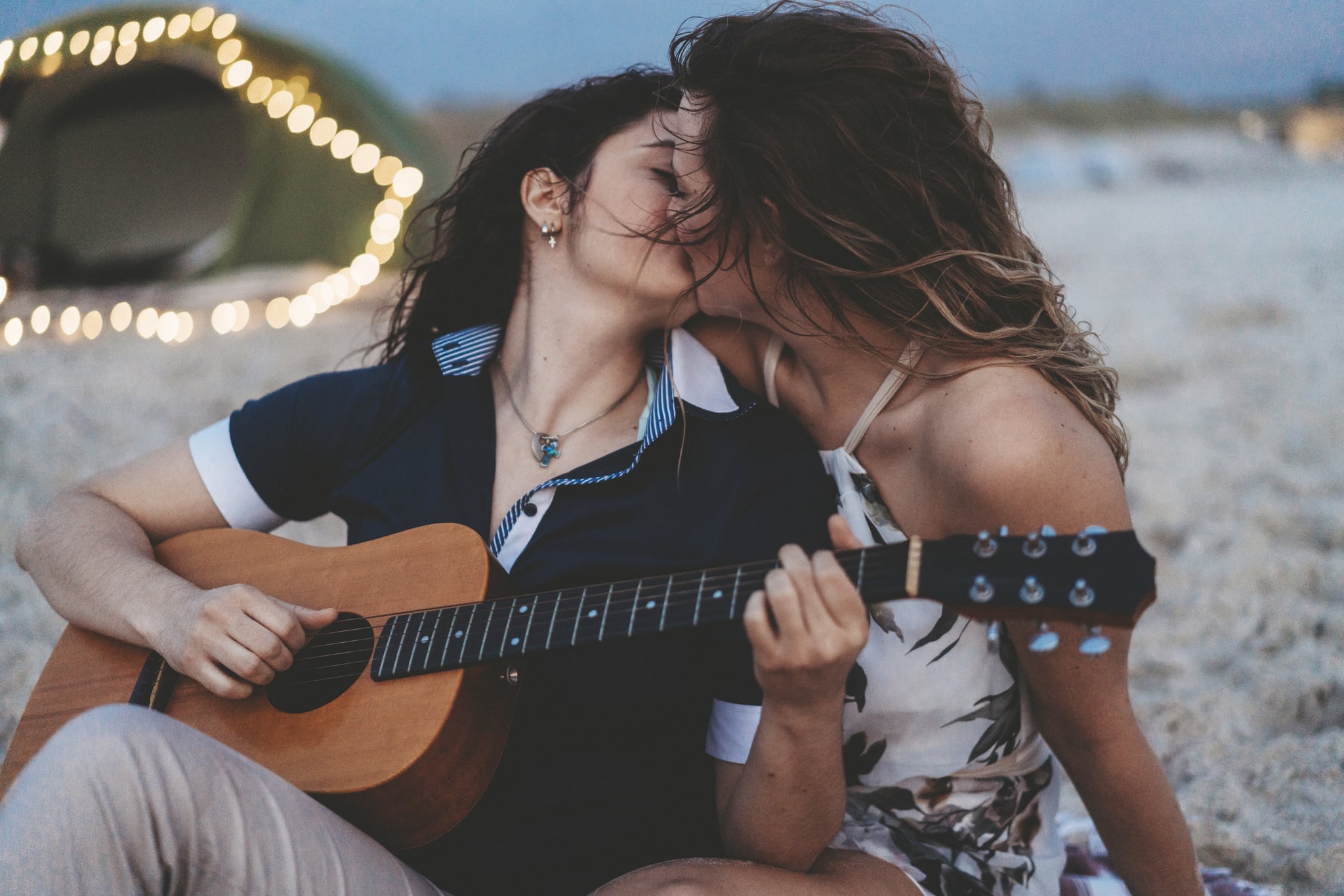 Young adult couple playing guitar at sunset near a tent on the beach