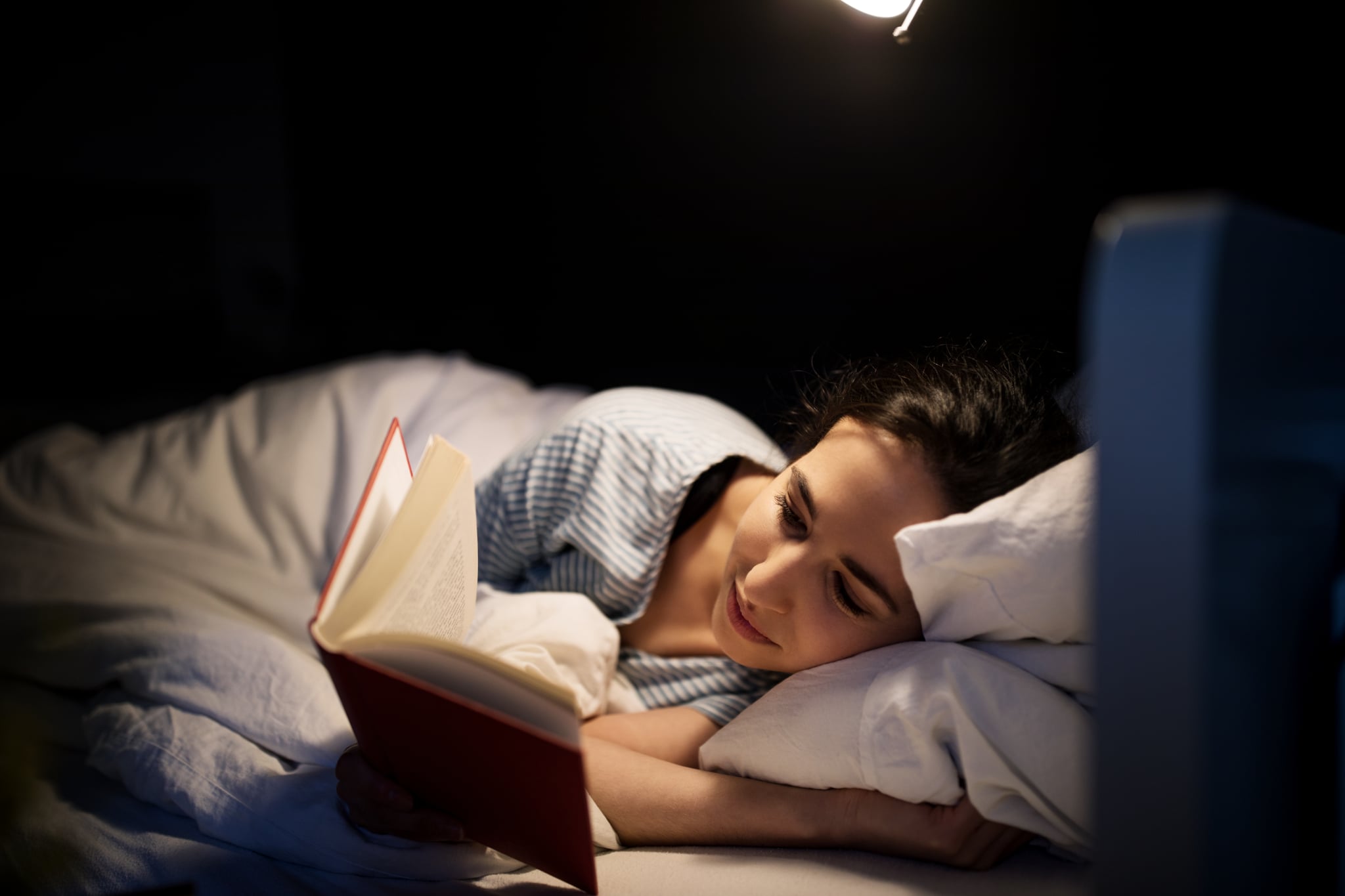 Young woman lying in her bed reading a book at night