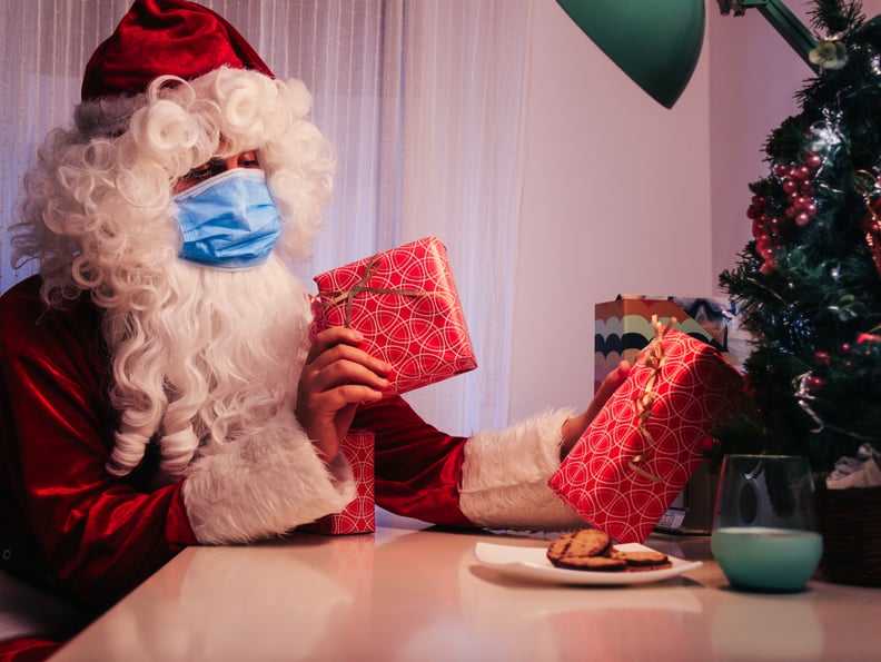 santa claus inside a house with a blue face mask, a glass of milk and cookies putting gifts on christmas tree