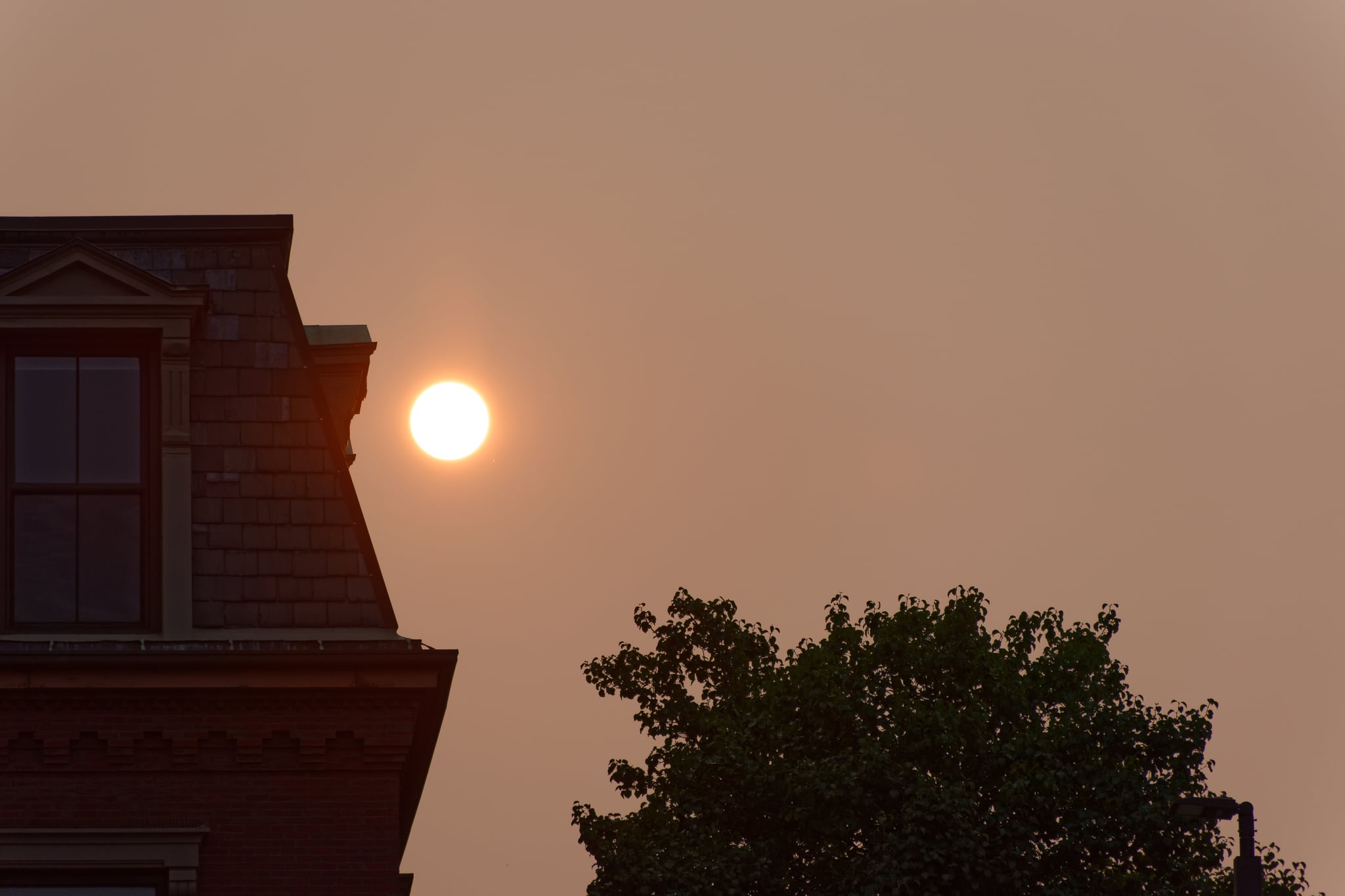 Smoke from the distant wildfires in the West turns Boston's sunrise red and hazy. The sun has cleared the horizon but is low enough to be partially framed by the silhouettes of a residential building and tree.