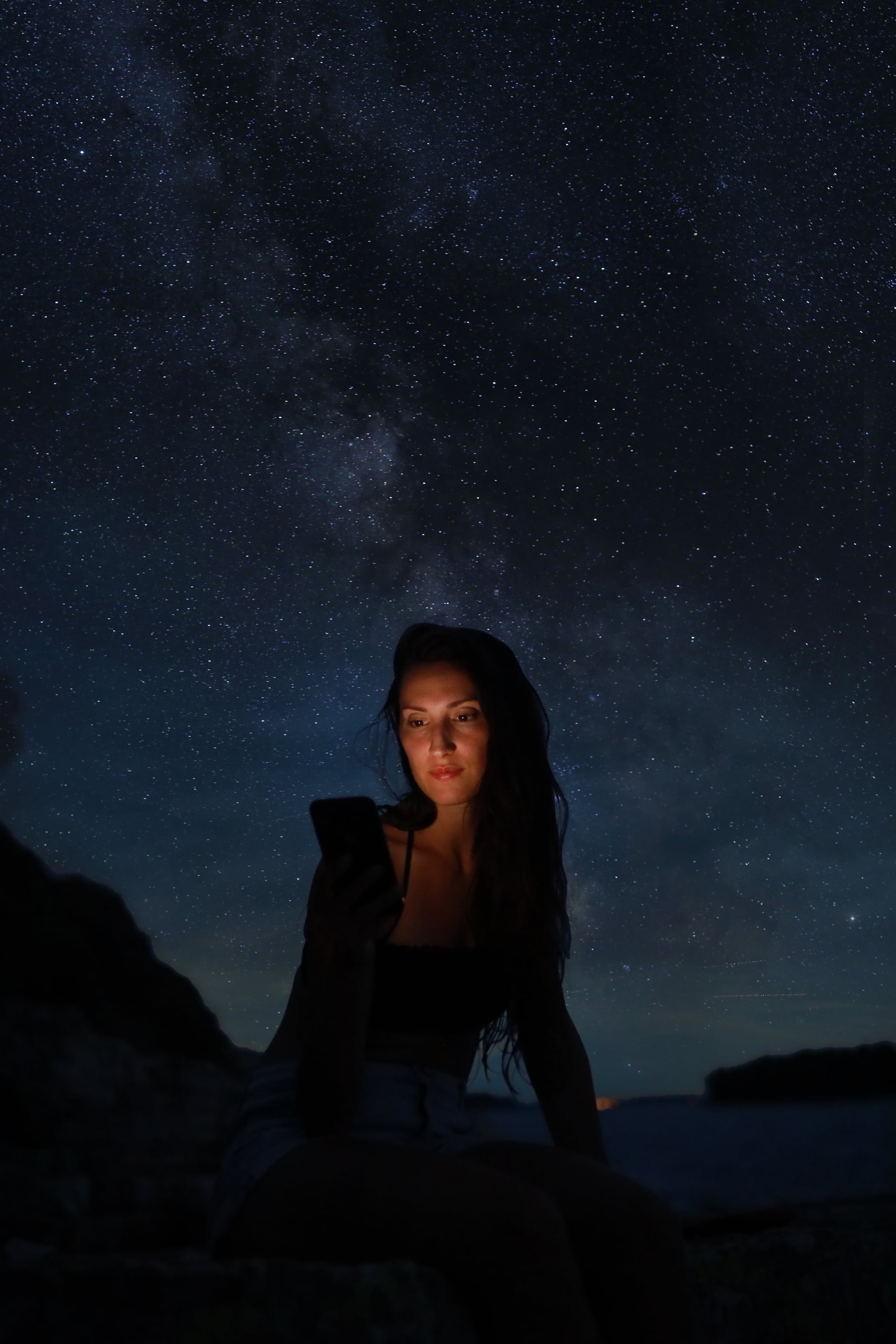 Woman sitting on the beach under the starry sky, looking at her cellphone