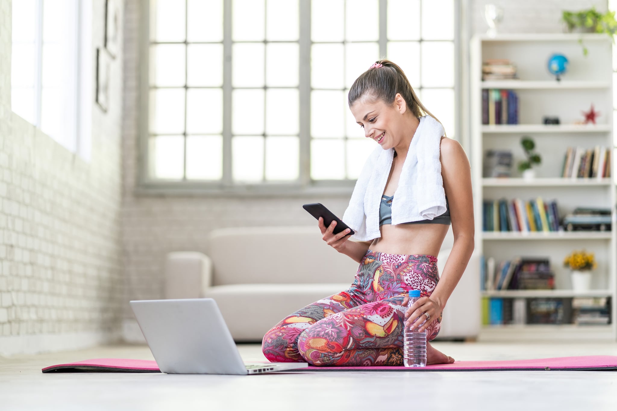 Woman using smart phone on exercise mat in front of her laptop