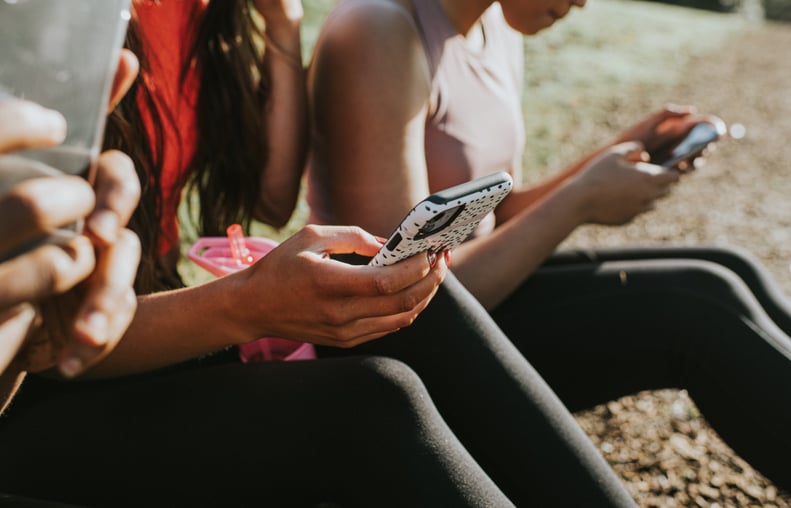 Three woman sit outside in the sun and hold their smart phones. They all look at their screens, while using fingers and thumbs to type. Conceptual with space for copy.