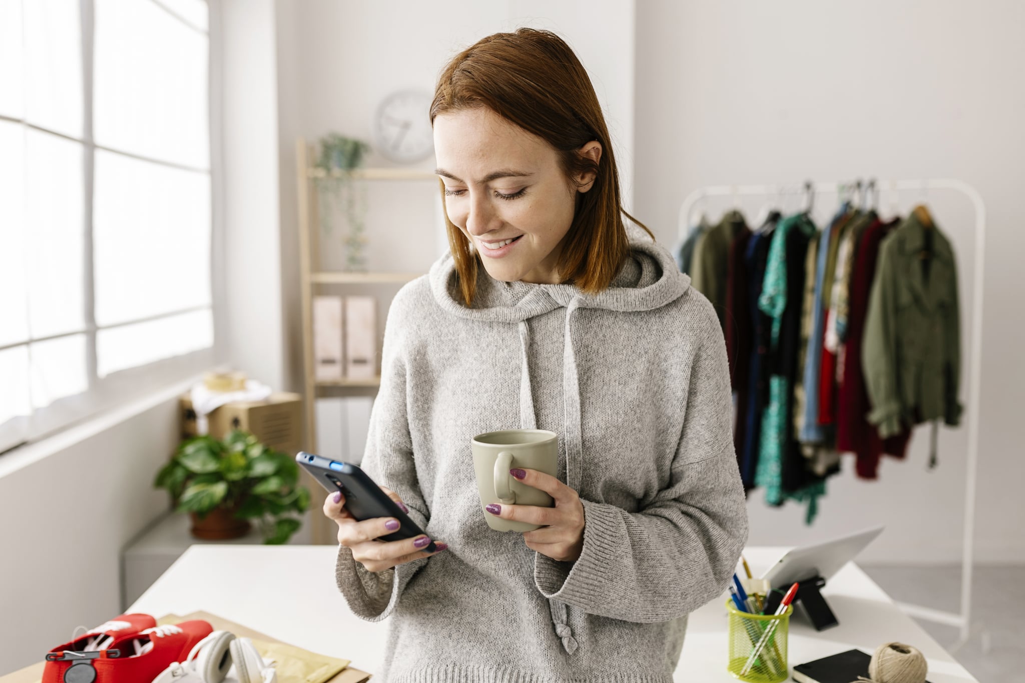 Female owner using cell phone and drinking coffee while standing in home office - Freelancer worker and small business concept