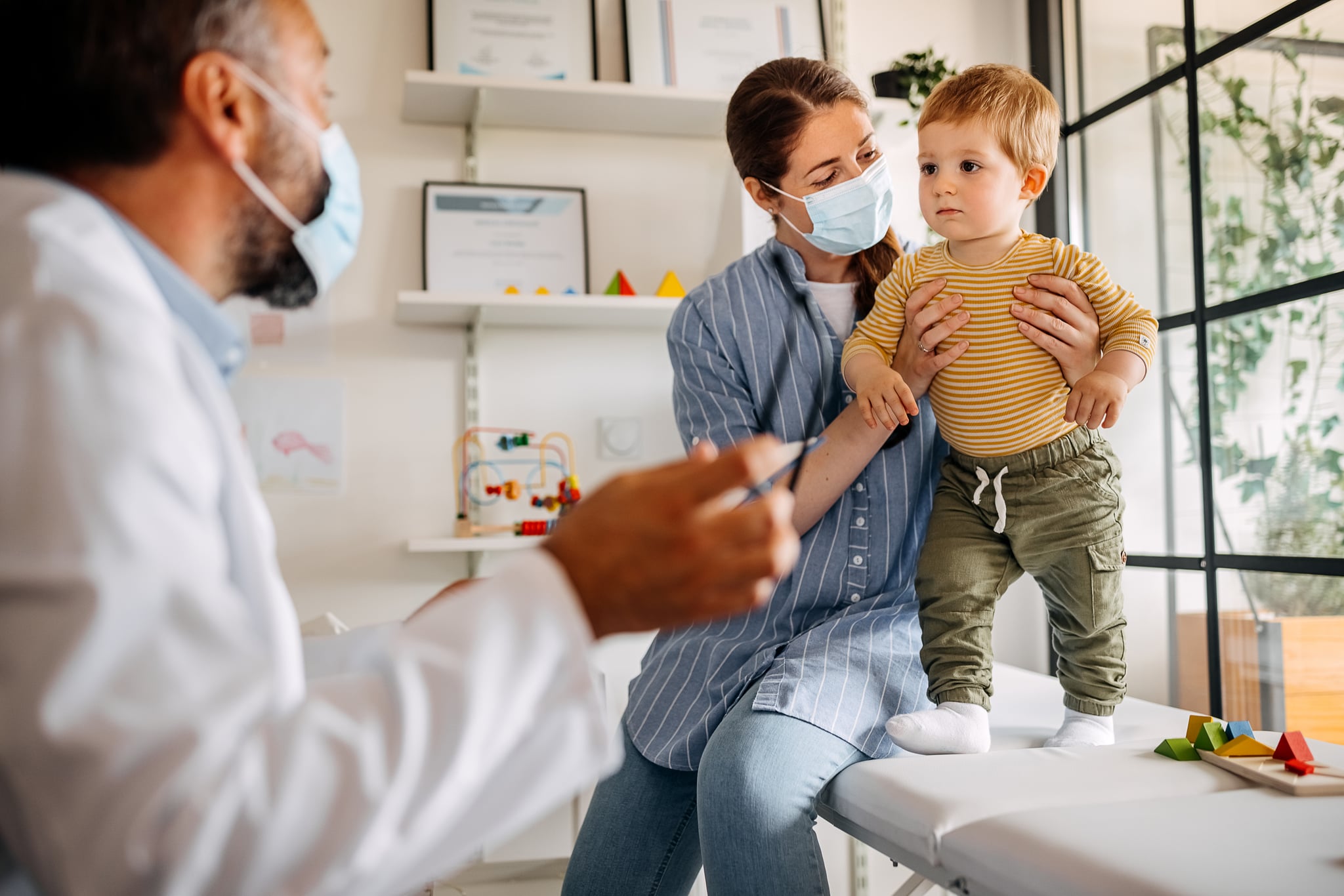 Cute little frightened baby boy standing with young and beautiful mother wearing face protection mask at medical clinic with senior pediatrician doctor