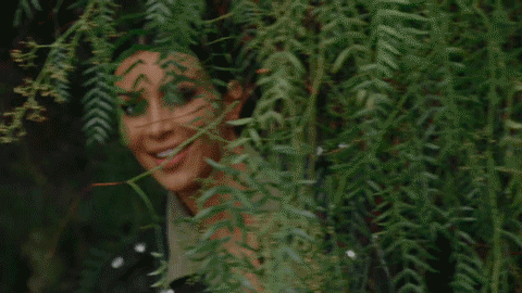 Kim Kardashian lurks behind a bush. She looks like she's sneaking or eavesdropping.