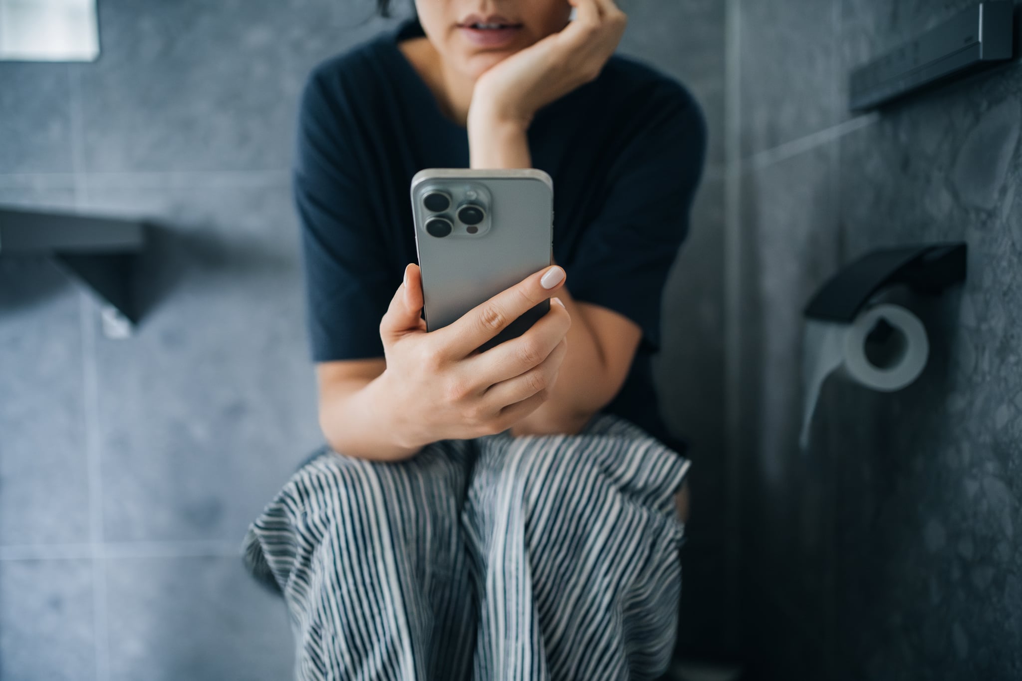 Cropped shot of young woman using phone while sitting on toilet in bathroom