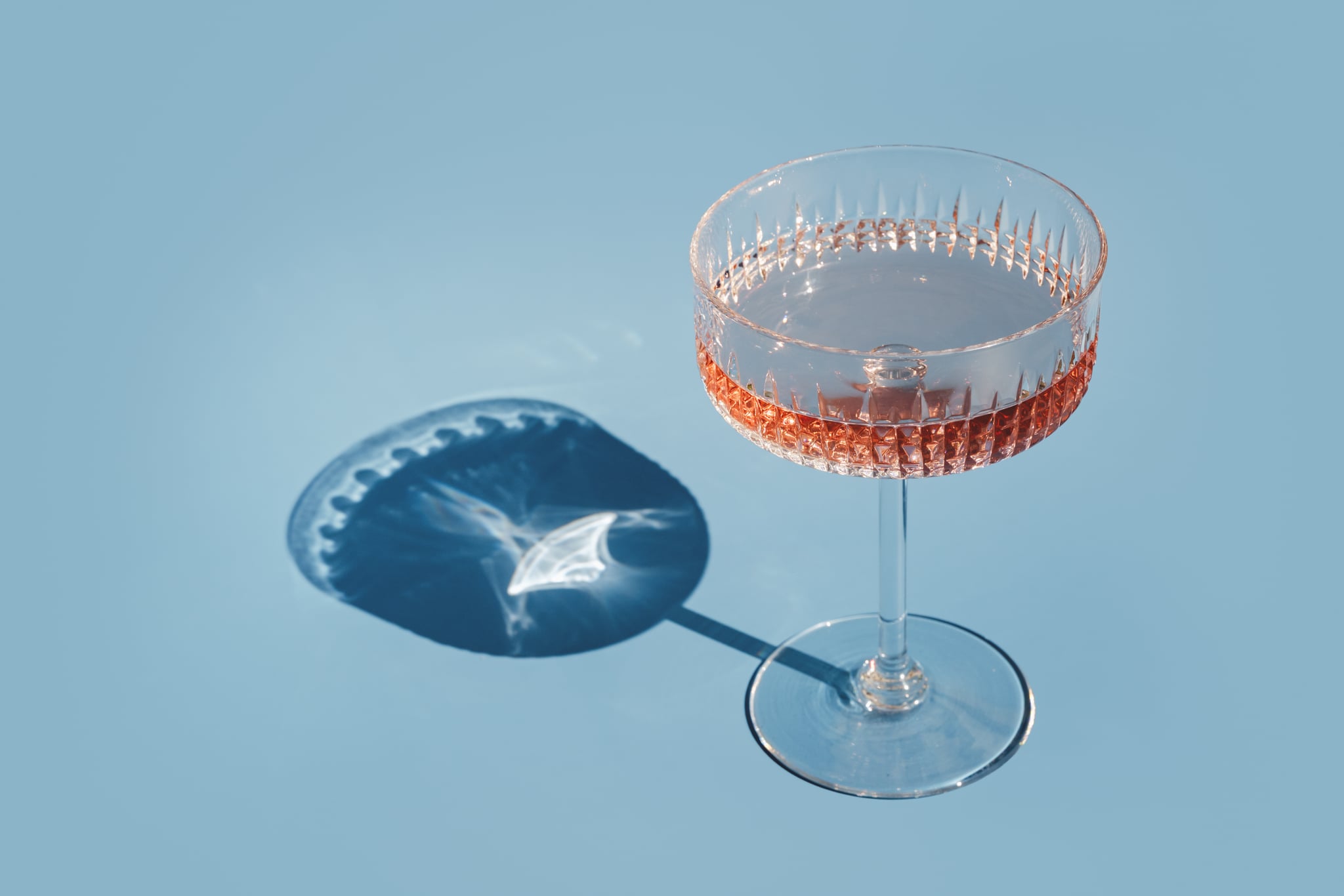 Rose wine refreshing summer drink in crystal champagne saucer glass on sunny day with shadows. Garden party cocktail and pink mocktail. blue tablecloth Elevated view. Sparkling blush wine