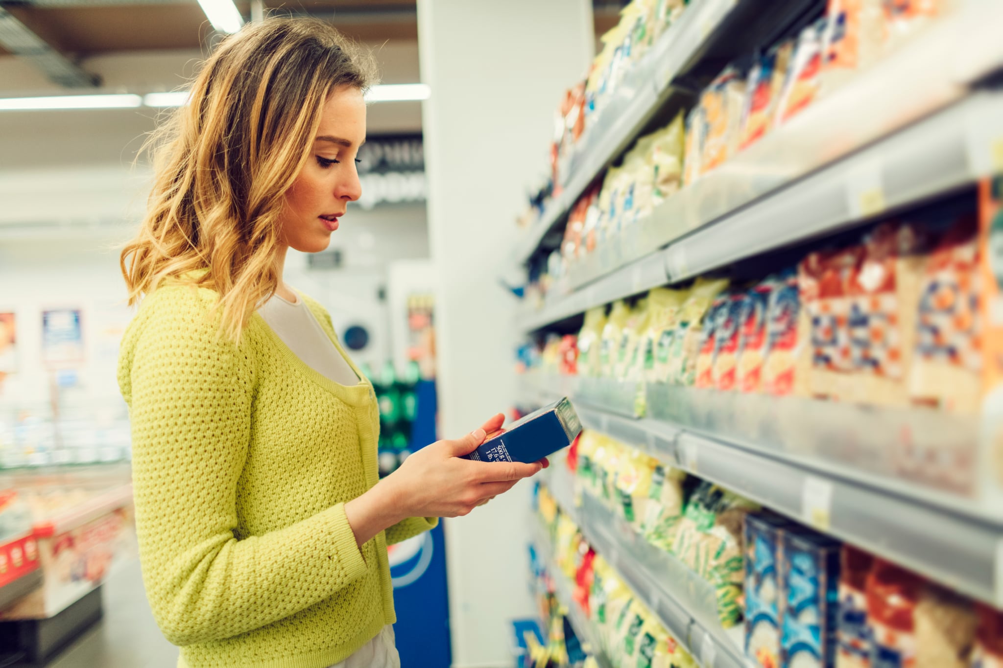 Young smiling woman groceries shopping in local supermarket. She is standing, holding box and reading nutrition facts.