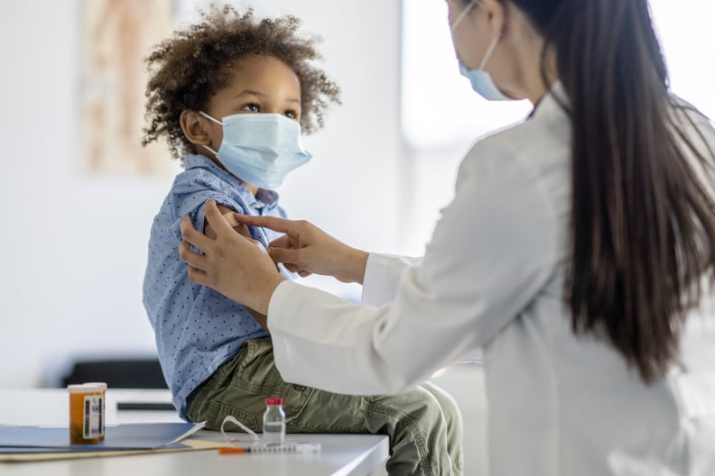 一个可爱的小混血男孩坐在检查台上上收到一个免疫。他抬头,微笑着医生把他手臂上的绷带。都是保护他们免受COVID带医用口罩。