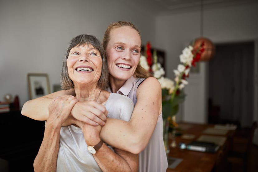 Cheerful mature woman embracing senior mother at home and looking away. Portrait of elderly mother and middle aged daughter smiling together.