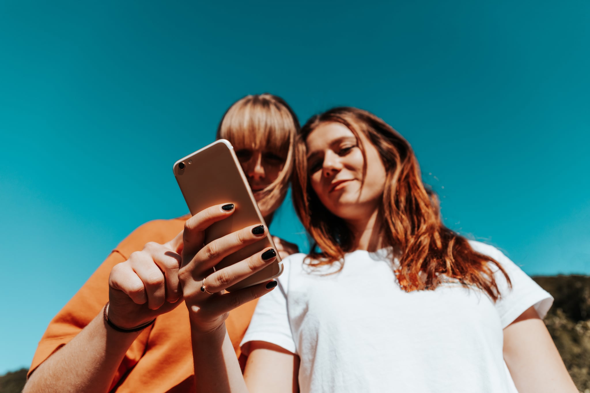 Young woman standing side by side reading the social media posts, news and messages on a mobile phone together. Selective Focus. Shot from below against the summer sky. Youth Culture Summer Lifestyle Concept.
