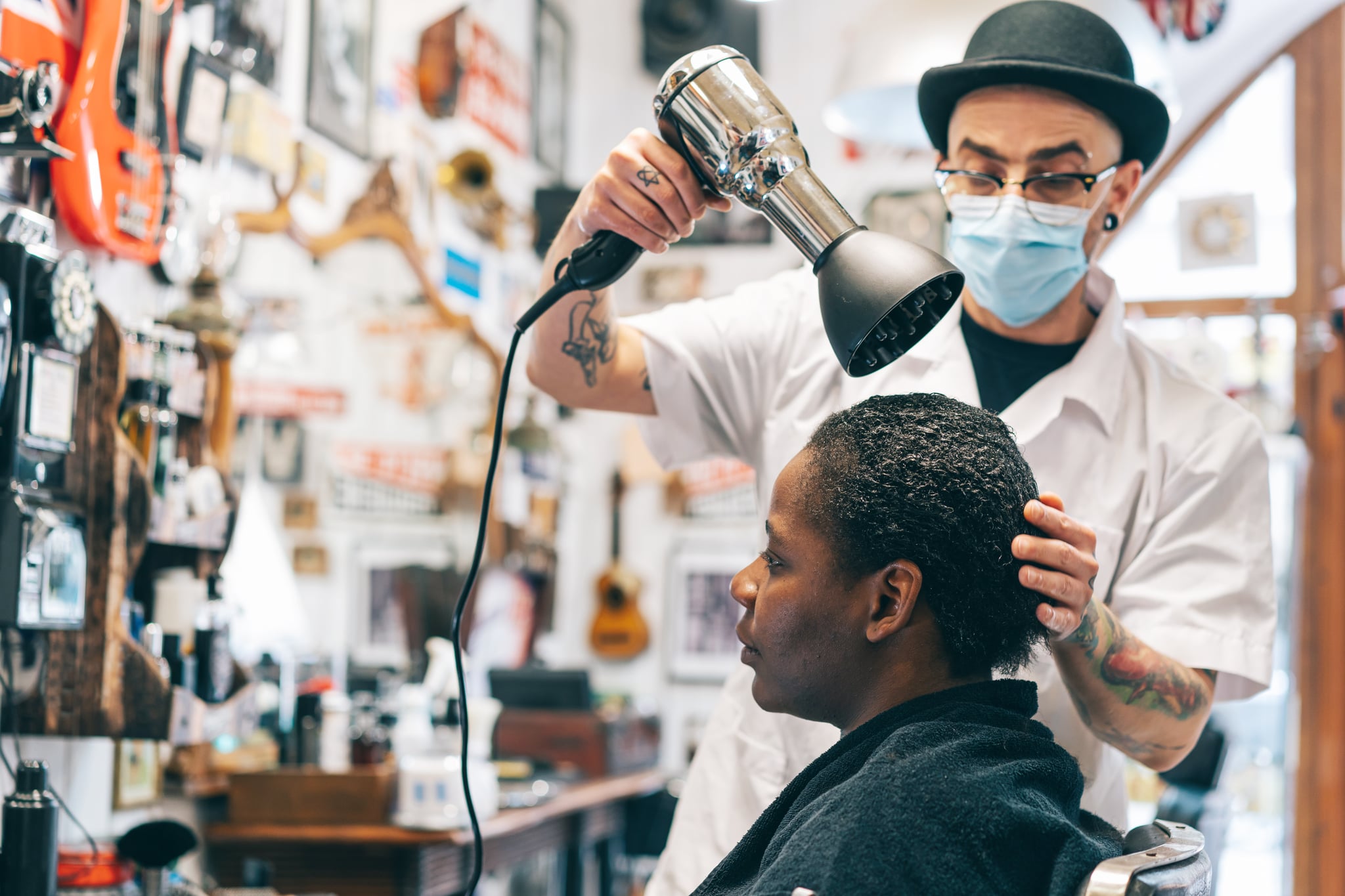 Male hairdresser wearing protective face mask and drying afro woman's hair with hair dryer in beauty studio