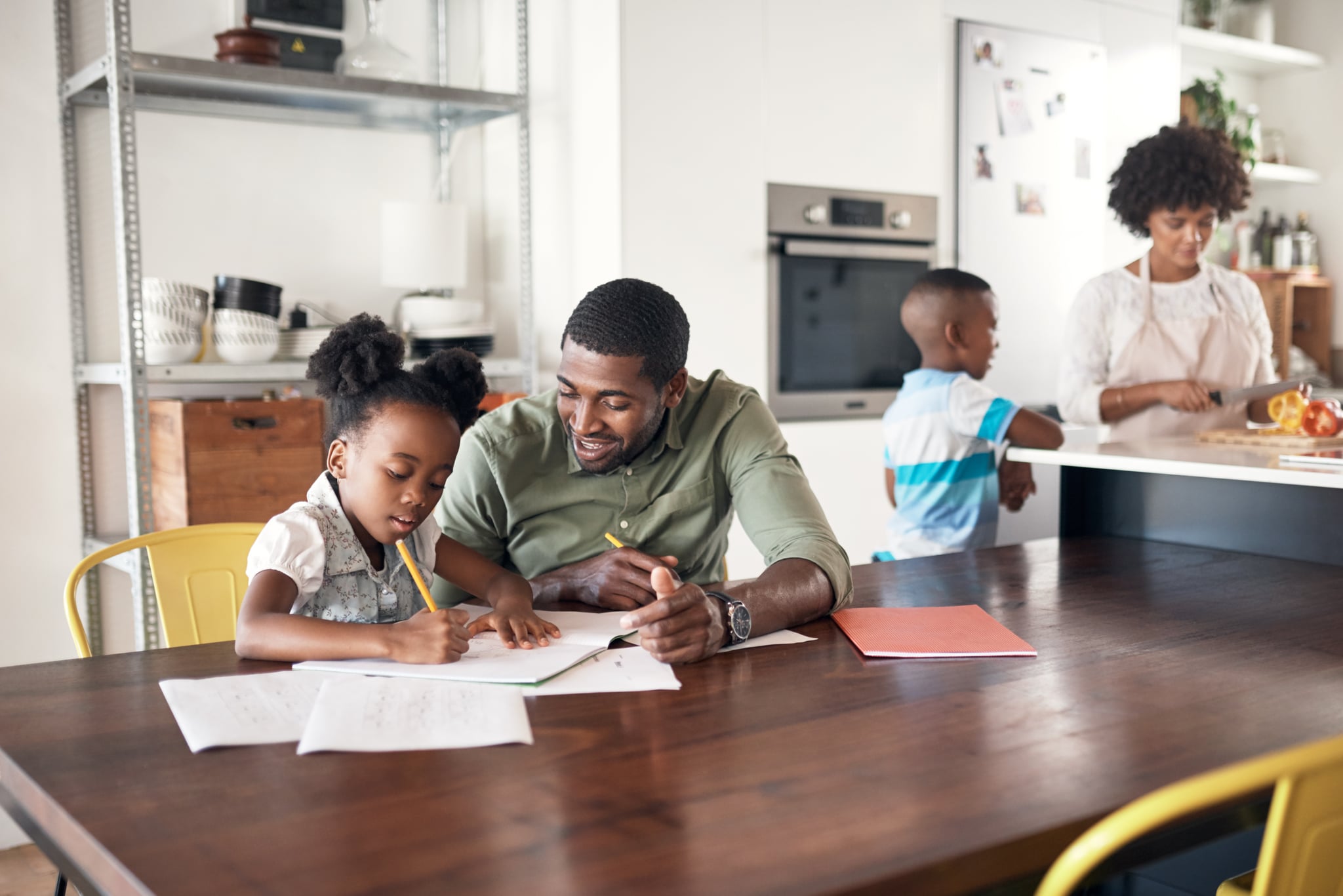 Shot of young man helping his daughter with her homework while her mother cooks in the background
