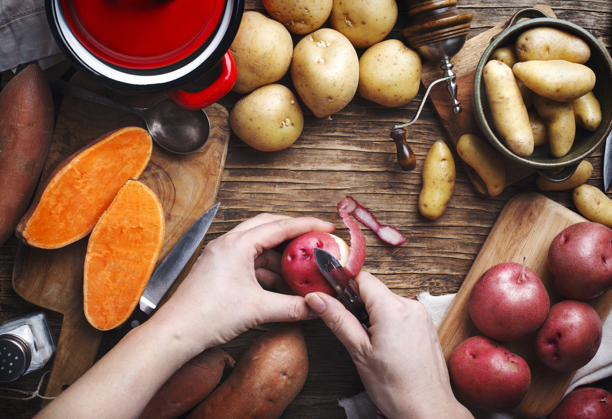 Womans hand peeling a potato, top view. Variety of raw uncooked organic potatoes: red, white, sweet  and fingers potatoes over wooden background.