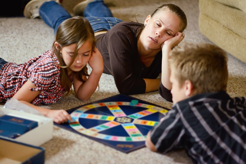 A mother nods off to sleep as her two children go on playing a board game.