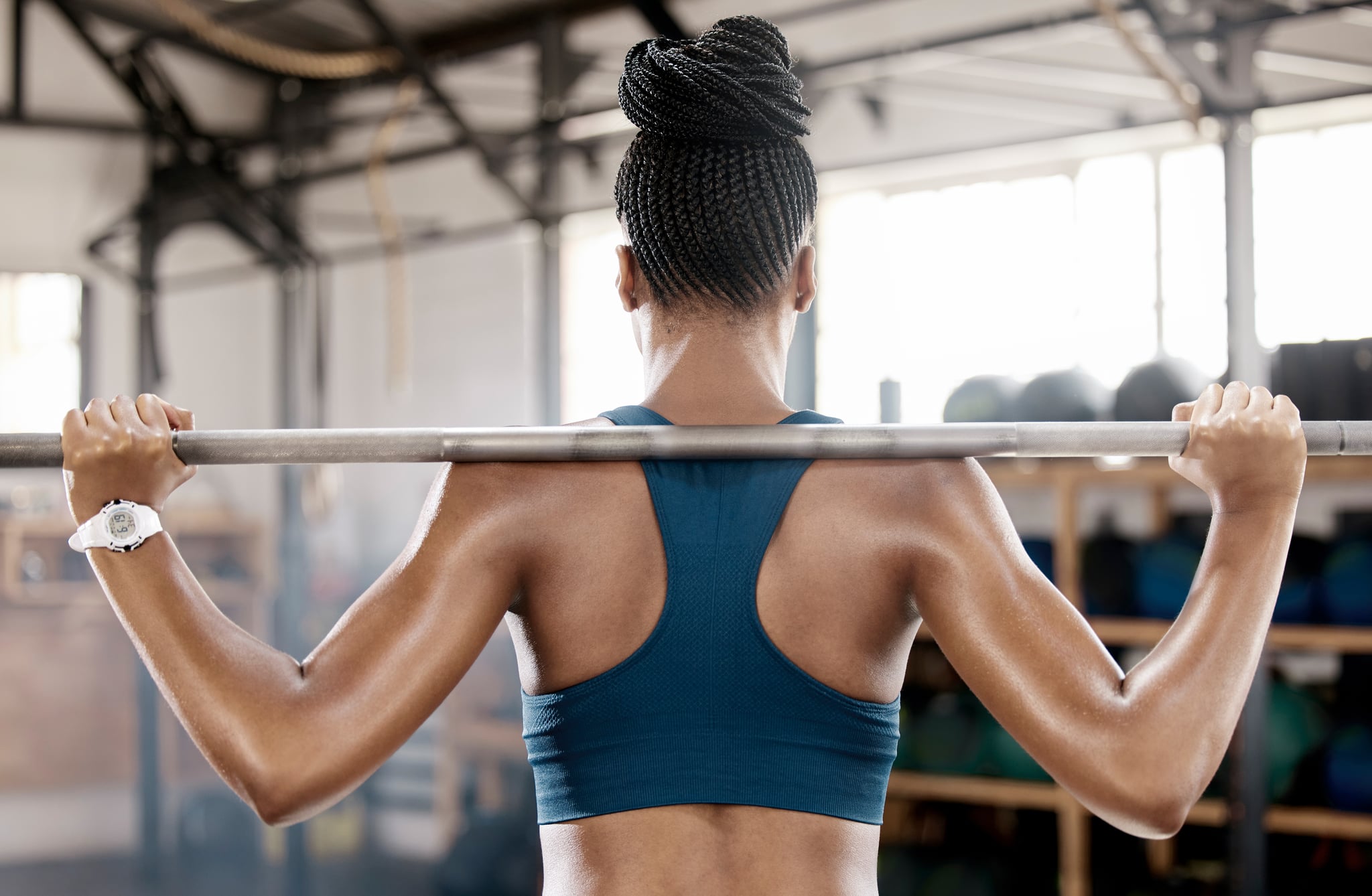 Back view of Black woman holding a barbell across her shoulders in the lower position of a press exercise.